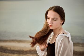 Young woman stands contemplatively by serene seaside, with gentle waves in background. Her relaxed...