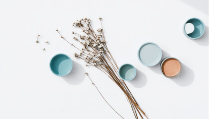 Minimalist flat lay of dried flowers and pastel-colored cups on a white background with subtle shadows and textures