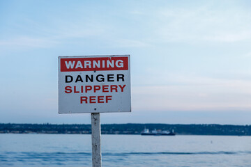 Warning sign indicating danger and slippery reef near a calm waterfront, with a ship visible in the...