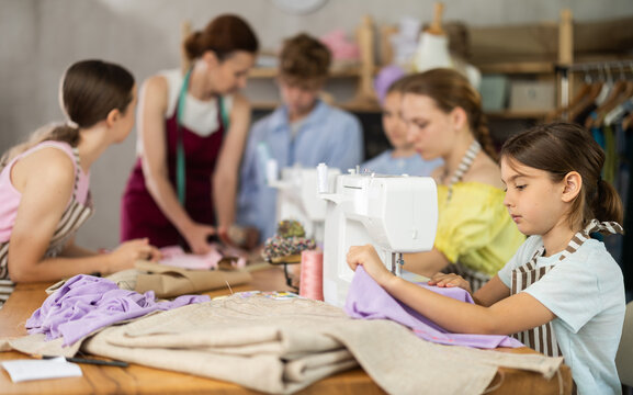 Girl child sews during practical lesson at school of young fashion designers and tailors. Classmates are doing preparatory work together in background.