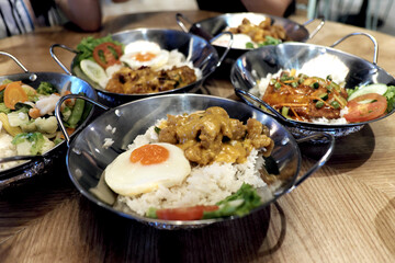 Creamy salted egg chicken with a sunny-side up egg over rice, served in a sizzling pan. The background is blurred with bokeh, showing hints of other tables and diners, indicating a busy but relaxed 