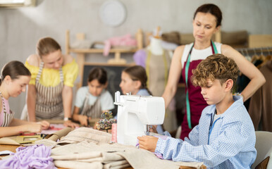 Cheerful boy working at sewing machine during childrens class in cozy school workshop while female...