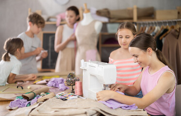 Teenage girl works on sewing machine while teacher and students learn how to cut fabric