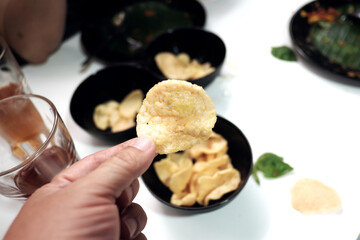 A hand holds up a crispy prawn cracker, with more bowls of crackers on a white table.