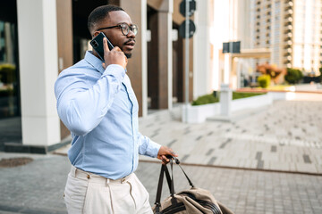 Black businessman walking outside while on the phone carrying a bag in an urban setting during...
