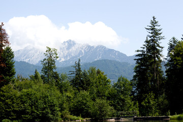 Lush green forest surrounds a calm lake with majestic, cloudy mountains in the background under a...