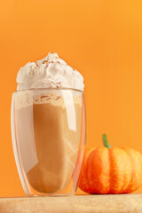 Homemade sweet pumpkin spice latte decorated with whipped cream topping served in drinking glass on wooden table with pumpkin against bright orange background as cozy autumn hot coffee festive drink 