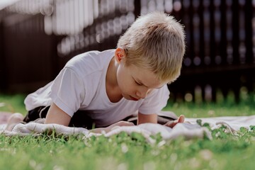 A boy with hearing aids plays on the grass while engaging with gadgets - drawing on a tablet and...