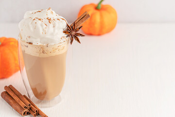 Sweet cozy pumpkin spice latte decorated with with whipped cream topping and anise and cinnamon stick garnish on white wooden table with copy space served in glass as autumn hot coffee festive drink