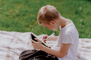 A boy with hearing aids plays on the grass while engaging with gadgets - drawing on a tablet and...