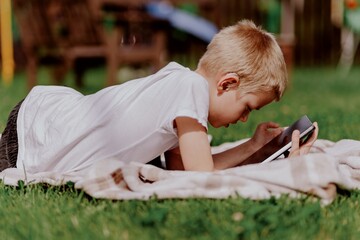 A boy with hearing aids plays on the grass while engaging with gadgets - drawing on a tablet and...