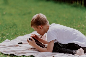A boy with hearing aids plays on the grass while engaging with gadgets - drawing on a tablet and...