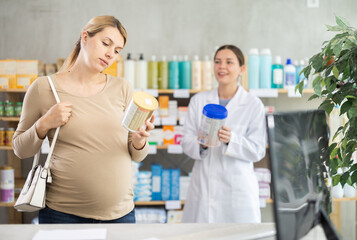 Fototapeta premium Caring expecting mother reading nutritional label on baby formula to ensure quality and safety, while smiling pharmacist presenting other option in background