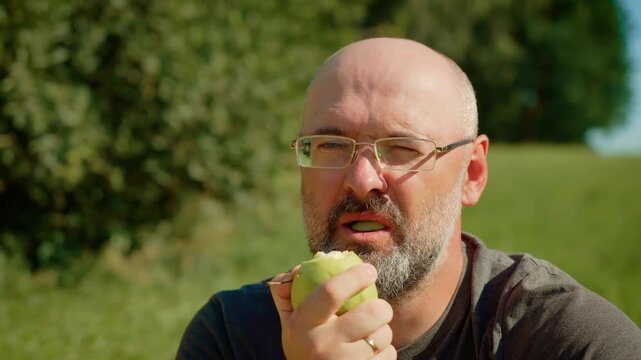 Bald man with glasses eating green apple, biting into crisp texture. Male chewing fresh green apple, enjoying tart flavor. Guy savoring juicy green apple outdoors, tasting refreshing sweetness