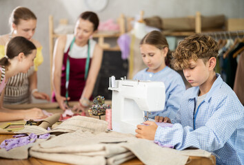 Teen male student sews product using sewing machine. Pupil are actively cooperating with each other...