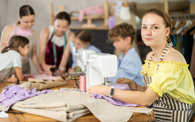Engaged teenage schoolgirl enjoying group sewing class, stitching at machine in school tailoring...