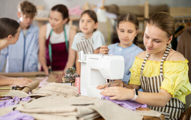 Teenage girl works on sewing machine while teacher and students learn how to cut fabric