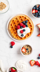 A close-up of a golden waffle topped with whipped cream, fresh blueberries, and strawberries on a white marble surface, surrounded by additional berries and breakfast items, creating an inviting and c