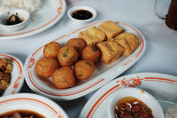 A table displaying various Indonesian dishes, with fried tofu and potato fritters (perkedel) as the centerpiece, set for a communal meal.