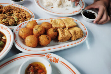 A table displaying various Indonesian dishes, with fried tofu and potato fritters (perkedel) as the centerpiece, set for a communal meal.