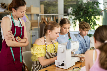 Engaged schoolgirl about fifteen practicing stitching at machine under guidance of female...