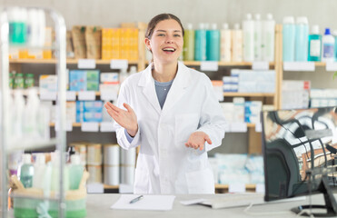 Friendly young female pharmacy worker in white coat gesturing with open hands, offering assistance...