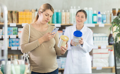 Fototapeta premium Pregnant woman in pharmacy scans QR code on infant formula product, studies product in detail, collects information online. Girl pharmacist offer similar product in background.
