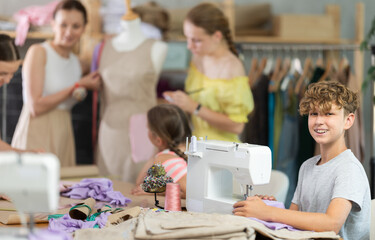 Teen boy child sews during practical lesson at school of young fashion designers and tailors. Classmates are doing preparatory work together in background.