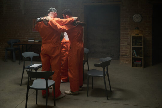 Group of four adult men standing in circle embracing each other in prison visiting room wearing orange jumpsuits, empty chairs surrounding them