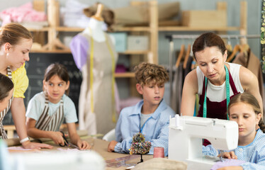 Positive friendly female sewing instructor guiding preteen schoolgirl stitching at machine during class while classmates working with fabric and patterns in creative workshop setting..