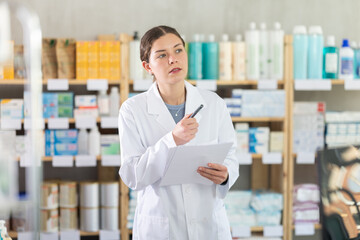 Focused young female apothecary taking records of stock using paper in chemistry