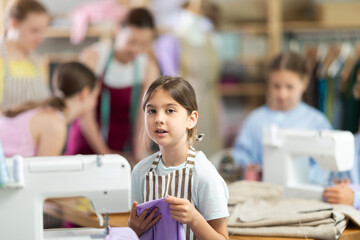 Girl child with cloth in hands examines product, school of young fashion designers. Students in...