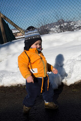 An adorable toddler dressed in a warm orange jacket and a striped hat stands in a snowy landscape,...
