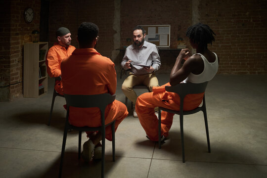 Multiethnic group of young adult men sitting in circle participating in group therapy session in prison setting with male facilitator holding clipboard leading discussion