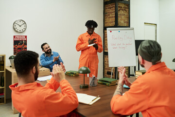 Black teenage boy standing and reciting poem in front of group of men wearing orange uniforms, men sitting at table clapping, whiteboard displaying Poetry Day