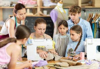 Focused teenage girl focusing on sewing at machine during class while female teacher correcting...