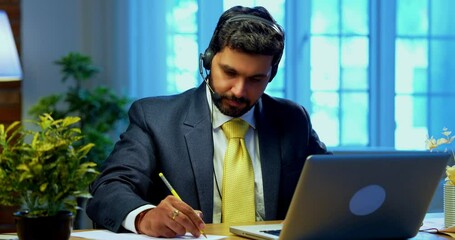Taking notes in video call on laptop at office desk, Indian Asian businessman with headphones focused on virtual meeting, corporate executive engaged in remote work, digital communication - Powered by Adobe