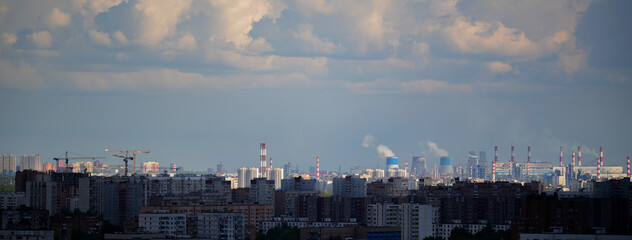 Obraz premium Cityscape showing a mix of residential buildings, construction sites with tower cranes, and a distant industrial zone with chimneys and cooling towers emitting steam. Concept of urban development, cit