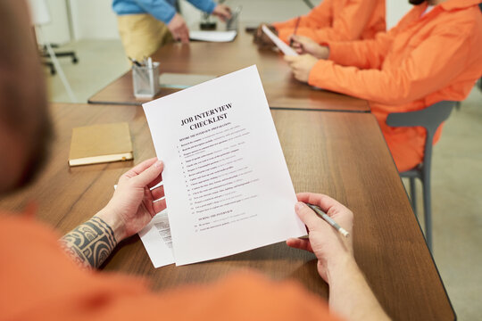 Caucasian man with tattooed arm holding job interview checklist, while sitting at table with other inmates in orange uniforms during educational program in prison classroom