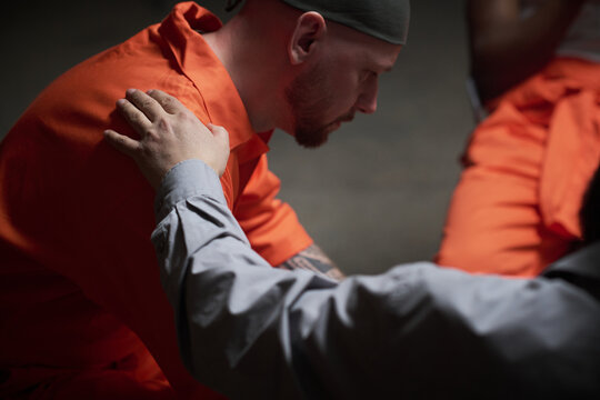 Caucasian man wearing prison uniform sitting with head bowed receiving comforting gesture from therapist during group session in correctional facility