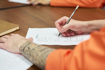Caucasian young adult man with tattooed arm filling out resume form at desk during prison education program, another inmate visible in background participating in learning activity