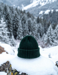 Cozy green knitted beanie rests on a snowcovered rock, evergreen trees blurred in the background. Winter fashion concept, for lifestyle or outdoor adventure themes.