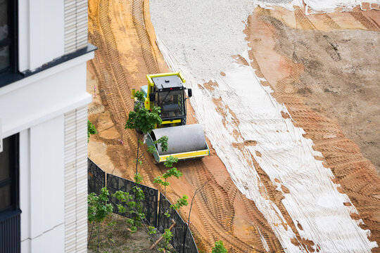Top-down view of a construction site where a road roller machine is compacting soil and gravel layers over geotextile fabric. The image captures the organized progress of groundwork preparation