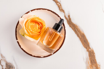 A chic bottle with a dropper of a cosmetic beauty care product lies on a plate with a rose head and a marble abstract background. natural cosmetics.