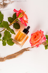 A natural beauty product in a clear dropper bottle on a marble abstract background among pink roses. vertical top view. A blank bottle mockup.
