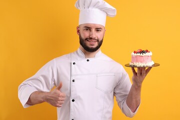 Happy confectioner in uniform holding cake with berries and showing thumbs up on orange background
