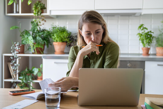 Pensive sorrowful woman reviewing bills with laptop and glass of water in home kitchen. Financial stress, debt issues, utility pay, household budget finance struggles, overdue accounts, money problems - Powered by Adobe