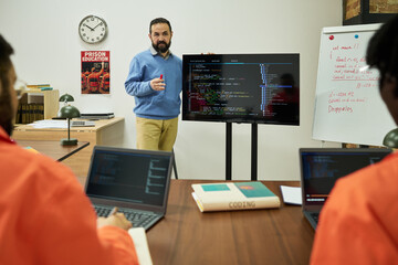 Middle aged Caucasian man explaining computer programming code on large monitor to group of young...