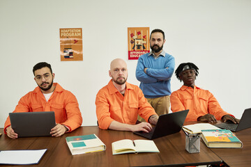 Three young adult men sitting at table using laptops with notebooks open, middle aged Caucasian man...