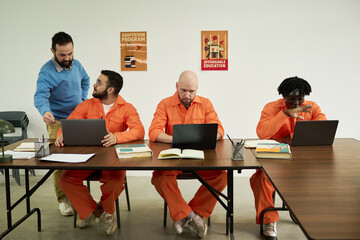 Middle aged Caucasian man assisting three adult men in orange uniforms working on laptops at table,...